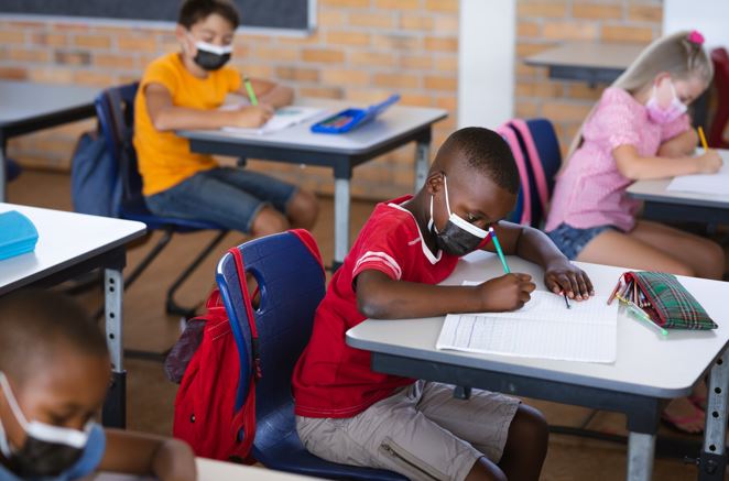 African american boy wearing face mask studying while sitting on his desk in the class at school