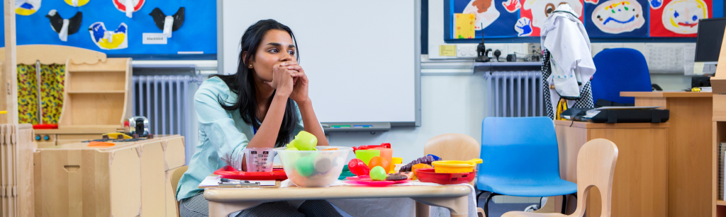 Young female teacher sitting alone in classroom at children's desk with a discouraged demeanor 